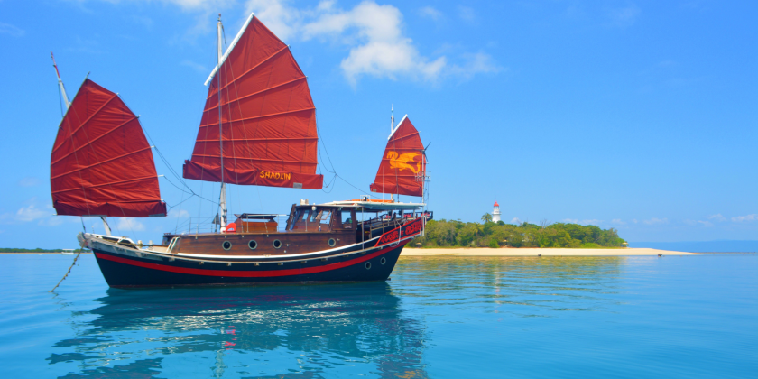 Boat with red sails near a sandy beach and distant lighthouse on a sunny day.